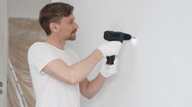 Beyaz tişört ve koruyucu eldiven giyen orta yaşlı erkek işçi, ev yenileme çalışmaları sırasında kablosuz matkapla duvarda delik açıyor. Dikey görünüm.