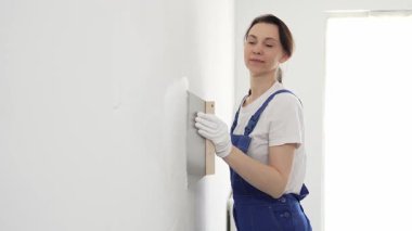 Koruyucu eldivenler ve mavi inşaat tulumları giyen kadın inşaat işçisi, tadilat projesi sırasında profesyonel alçı levha bantlı bıçak kullanarak duvarlara sorunsuzca alçı yayıyor. Portre