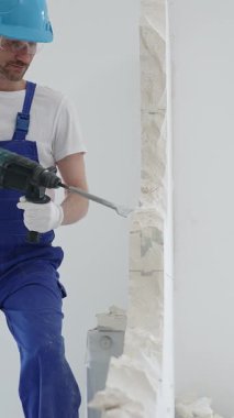 Mavi tulumlu ve koruyucu beyaz eldivenli bir inşaat işçisi, beyaz duvarı döner çekiç matkabıyla yıkıyor, toz üretiyor, yatay görüşü kapatıyor. Yenileme konsepti.