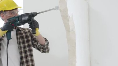 Bej kareli gömlek, sarı başlık ve koruyucu eldiven giyen inşaat işçisi, döner çekiç matkabıyla beyaz duvarı yıkıyor, toz ve portre görüntüsü yaratıyor. Yenileme konsepti.