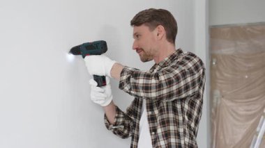 İşaretli gömlek ve beyaz eldiven giyen orta yaşlı bir adam, ev yenileme çalışmaları sırasında kablosuz matkapla duvarda delik açıyor. Dikey görünüm.