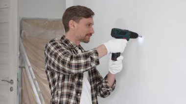İşaretli gömlek ve beyaz eldiven giyen orta yaşlı bir adam, ev yenileme çalışmaları sırasında kablosuz matkapla duvarda delik açıyor. Dikey görünüm.