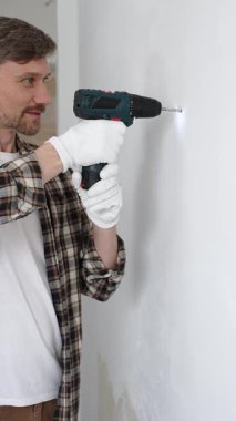 Mavi üniformalı, güvenlik gözlüğü ve eldiven takan profesyonel kadın işçi, tadilat sırasında beyaz duvarda delik açmak için kablosuz matkap kullanıyor. Dikey görünüm.