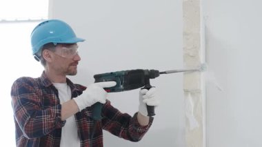 Kırmızı kareli gömlek, mavi başlık ve koruyucu eldiven giyen inşaat işçisi, döner çekiç matkabıyla beyaz duvarı yıkıyor, toz ve portre görüntüsü yaratıyor. Yenileme konsepti.