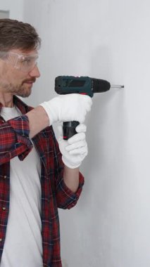 Kırmızı tişört ve beyaz eldiven giyen orta yaşlı erkek işçi, ev yenileme çalışmaları sırasında kablosuz matkapla duvarda delik açıyor. Dikey görünüm.