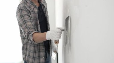 Eldiven ve ekose gömlek giyen kimliği belirsiz erkek inşaat işçisi, yenileme projesi sırasında pürüzsüz bir yüzey oluşturmak için beyaz bir duvar alçılamak için son malayı kullanıyor..