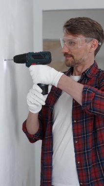 Kırmızı tişört ve beyaz eldiven giyen orta yaşlı erkek işçi, ev yenileme çalışmaları sırasında kablosuz matkapla duvarda delik açıyor. Dikey görünüm.