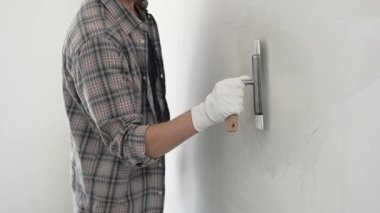 Eldiven ve ekose gömlek giyen kimliği belirsiz erkek inşaat işçisi, yenileme projesi sırasında pürüzsüz bir yüzey oluşturmak için beyaz bir duvar alçılamak için son malayı kullanıyor..