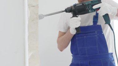 Mavi tulumlu ve koruyucu beyaz eldivenli tanımlanamayan erkek inşaat işçisi, beyaz duvarı döner çekiç matkapıyla yıkıyor, toz üretiyor ve yatay görüşü kapatıyor. Yenileme