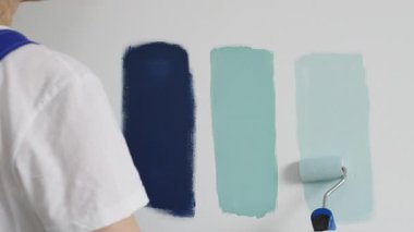 Eldiven giyen ressam beyaz bir duvara boya silindiri uyguluyor, mavi rengin çeşitli tonlarını test ederek iç tasarım için güzel bir eğim etkisi yaratıyor.