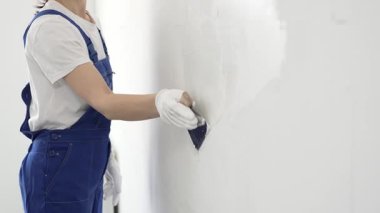 Meçhul inşaat işçisi beyaz eldiven ve mavi tulum giyen kadın, macun bıçağı kullanarak beyaz bir duvara alçı sürüyor. Ev yenileme projesi sırasında pürüzsüz bir yüzey oluşturuyor.