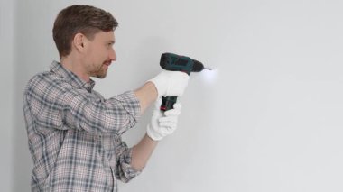 Gri gömlekli ve beyaz eldivenli orta yaşlı işçi, ev yenileme çalışmaları sırasında kablosuz matkapla duvarda delik açıyor. Dikey görünüm.
