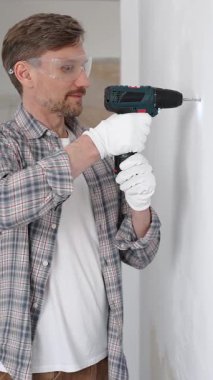 Gri gömlekli ve beyaz eldivenli orta yaşlı işçi, ev yenileme çalışmaları sırasında kablosuz matkapla duvarda delik açıyor. Dikey görünüm.