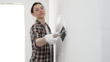 Koruyucu eldiven ve kareli gömlek giyen kadın, iç dekorasyon çalışmalarını boyamadan önce hasarlı bölgeleri yumuşatmak için beyaz duvar yüzeyine macun sürüyor..