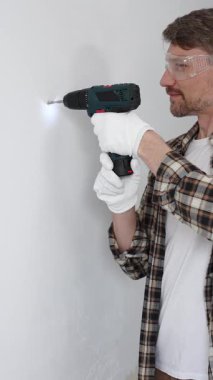 İşaretli gömlek ve beyaz eldiven giyen orta yaşlı bir adam, ev yenileme çalışmaları sırasında kablosuz matkapla duvarda delik açıyor. Dikey görünüm.