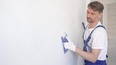 Koruyucu beyaz eldiven ve mavi inşaat tulumları giyen inşaat işçisi, iç dekorasyon projesi sırasında pürüzsüz bir yüzey hazırlamak üzere mavi el bloğuyla beyaz duvarı zımparalıyor..