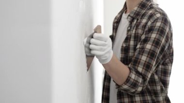 Bilinmeyen, koruyucu eldiven ve kareli gömlek giyen kadın inşaat işçisi, yenileme projesi sırasında beyaz duvara alçı yayıyor, macun bıçağıyla pürüzsüz yüzey.