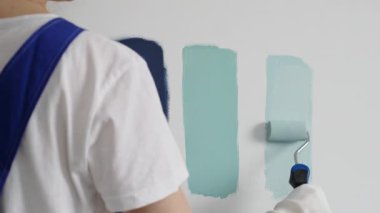 Eldiven giyen ressam beyaz bir duvara boya silindiri uyguluyor, mavi rengin çeşitli tonlarını test ederek iç tasarım için güzel bir eğim etkisi yaratıyor.