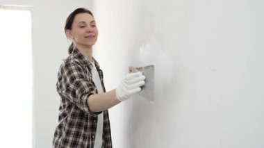 Koruyucu eldiven giyen ve kareli gömlekli bir kadın beyaz duvara macun yapıştırıyor, ev tadilatı sırasında spatulayla bileşik yayıyor, pratik ev geliştirme becerileri sergiliyor..