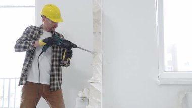 Bej kareli gömlek, sarı başlık ve koruyucu eldiven giyen inşaat işçisi, dönen çekiç matkabıyla beyaz duvarı yıkıyor ve orta uzunlukta toz üretiyor. Yenileme konsepti