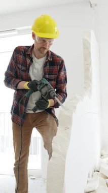 Bej kareli gömlek, sarı başlık ve koruyucu eldiven giyen inşaat işçisi, dönen çekiç matkabıyla beyaz duvarı yıkıyor ve orta uzunlukta toz üretiyor. Yenileme konsepti