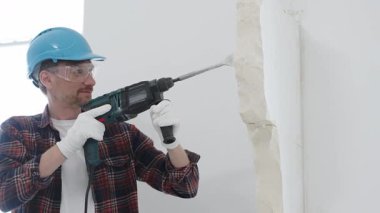 Kırmızı kareli gömlek, mavi başlık ve koruyucu eldiven giyen inşaat işçisi, döner çekiç matkabıyla beyaz duvarı yıkıyor, toz ve portre görüntüsü yaratıyor. Yenileme konsepti.