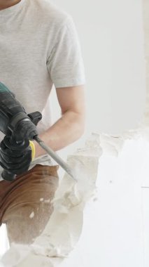 Beyaz tişörtlü ve koruyucu eldivenli, kimliği belirsiz bir inşaat işçisi, beyaz duvarı döner çekiç matkapıyla yıkıyor, yerde enkaz oluşturuyor, dikey yakın görüş açısında. Yenileme.