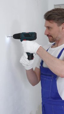 Mavi üniformalı profesyonel işçi, güvenlik eldivenleri, tadilat sırasında beyaz duvarda delik açmak için kablosuz matkap kullanıyor. Dikey portre görünümü.