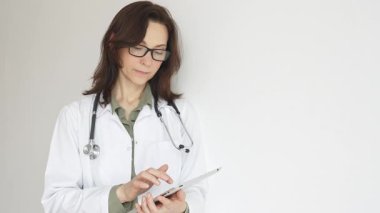 Yeşil bluz, steteskop ve gözlük üzerine beyaz önlük giyen doktor kadın, dijital tablet üzerindeki hasta kayıtlarını inceliyor. Tıbbi çalışma alanında doğal ışıklandırmayla duruyor. İlaç..