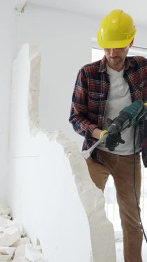 Bej kareli gömlek, sarı başlık ve koruyucu eldiven giyen inşaat işçisi, dönen çekiç matkabıyla beyaz duvarı yıkıyor ve orta uzunlukta toz üretiyor. Yenileme konsepti