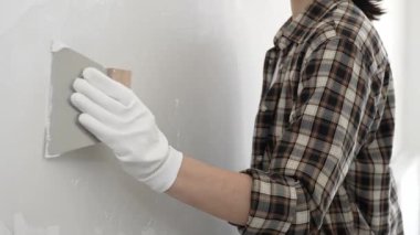 Bilinmeyen, koruyucu eldiven ve kareli gömlek giyen kadın inşaat işçisi, yenileme projesi sırasında beyaz duvara alçı yayıyor, macun bıçağıyla pürüzsüz yüzey.