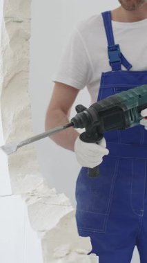 Mavi tulumlu ve koruyucu beyaz eldivenli tanımlanamayan erkek inşaat işçisi, beyaz duvarı döner çekiç matkapıyla yıkıyor, toz üretiyor ve yatay görüşü kapatıyor. Yenileme