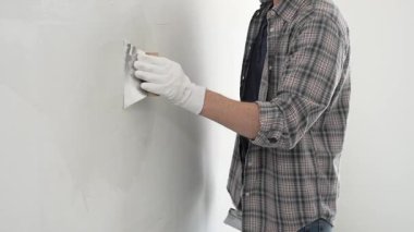 Eldiven ve ekose gömlek giyen tanınmamış inşaat işçisi, bir ev yenileme veya inşaat projesi sırasında beyaz bir duvara sıva uygulamak için macun bıçağı kullanıyor..