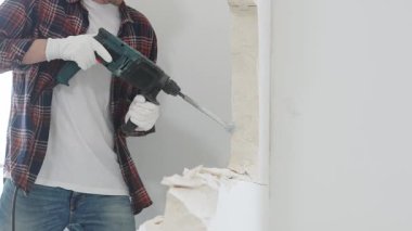 Kırmızı kareli kot pantolon ve koruyucu eldiven giyen kimliği belirsiz erkek inşaat işçisi, döner çekiç matkabıyla beyaz duvarı yıkıyor, toz üretiyor, yakın görüş sağlıyor. Yenileme konsepti.