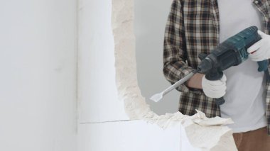 Tanınmayan bej kareli gömlek ve koruyucu eldivenler giyen erkek inşaat işçisinin yakın çekiç matkabıyla beyaz duvarı yıkıyor, toz üretiyor, yakın görüş sağlıyor. Yenileme