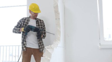 Bej kareli gömlek, sarı başlık ve koruyucu eldiven giyen inşaat işçisi, dönen çekiç matkabıyla beyaz duvarı yıkıyor ve orta uzunlukta toz üretiyor. Yenileme konsepti
