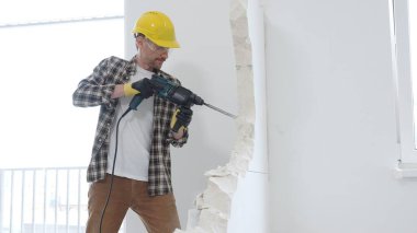 Bej kareli gömlek, sarı başlık ve koruyucu eldiven giyen inşaat işçisi, dönen çekiç matkabıyla beyaz duvarı yıkıyor ve orta uzunlukta toz üretiyor. Yenileme konsepti