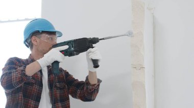 Kırmızı kareli gömlek, mavi başlık ve koruyucu eldiven giyen inşaat işçisi, döner çekiç matkabıyla beyaz duvarı yıkıyor, toz ve portre görüntüsü yaratıyor. Yenileme konsepti.