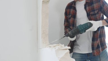 Kırmızı kareli kot pantolon ve koruyucu eldiven giyen kimliği belirsiz erkek inşaat işçisi, döner çekiç matkabıyla beyaz duvarı yıkıyor, toz üretiyor, yakın görüş sağlıyor. Yenileme konsepti.