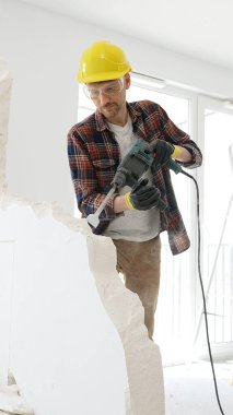 Kırmızı kareli gömlek giyen, sarı koruyucu kask takan erkek inşaat işçisi, beyaz duvarı döner çekiç matkabıyla yıkıyor, toz üretiyor, orta uzunlukta. Yenileme projesi.