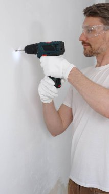 Beyaz tişört ve koruyucu eldiven giyen orta yaşlı erkek işçi, ev yenileme çalışmaları sırasında kablosuz matkapla duvarda delik açıyor. Dikey görünüm.
