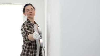 Kareli gömlek giyen kadın inşaat işçisi, beyaz duvar yüzeyini düzleştiriyor, profesyonel spatula tekniği ile macunu uyguluyor, iç dekorasyon konusunda yetenekli..