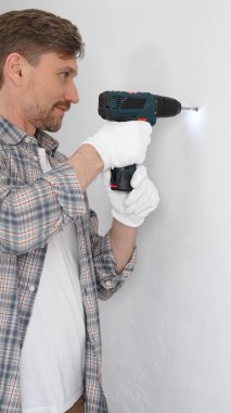 Gri gömlekli ve beyaz eldivenli orta yaşlı profesyonel işçi, ev yenileme çalışmaları sırasında kablosuz matkapla duvarda delik açıyor. Dikey portre görünümü.