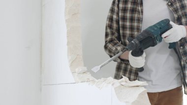 Tanınmayan bej kareli gömlek ve koruyucu eldivenler giyen erkek inşaat işçisinin yakın çekiç matkabıyla beyaz duvarı yıkıyor, toz üretiyor, yakın görüş sağlıyor. Yenileme