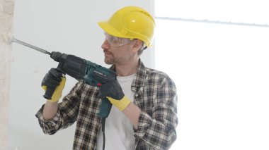 Bej kareli gömlek, sarı başlık ve koruyucu eldiven giyen inşaat işçisi, döner çekiç matkabıyla beyaz duvarı yıkıyor, toz ve portre görüntüsü yaratıyor. Yenileme konsepti.