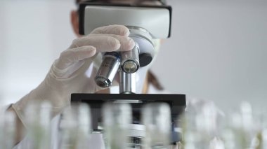 Beyaz koruyucu giysiler giyen kadın bilim adamı çağdaş araştırma ortamında test örneklerini mikroskop altında analiz ediyor. Tıp, sağlık ve bilim konsepti.