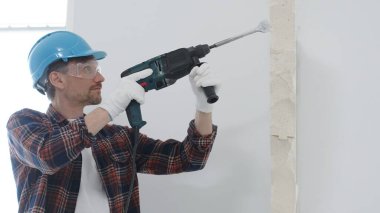 Kırmızı kareli gömlek, mavi koruyucu kask ve eldiven giyen erkek inşaat işçisi, döner çekiç matkabıyla beyaz duvarı yıkıyor, toz ve portre görüntüsü yaratıyor. Yenileme konsepti.