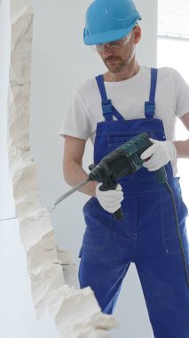 Mavi tulumlu ve koruyucu beyaz eldivenli bir inşaat işçisi, beyaz duvarı döner çekiç matkabıyla yıkıyor, toz üretiyor, yatay görüşü kapatıyor. Yenileme konsepti.