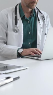 Doktor adam yeşil gömleğin üstüne beyaz önlük giyiyor ve steteskop da parlak bir tıbbi ofisteki dizüstü bilgisayarda yazarak sağlık ve teknolojinin kesişimini gösteriyor. İlaç ve sağlık hizmetleri.