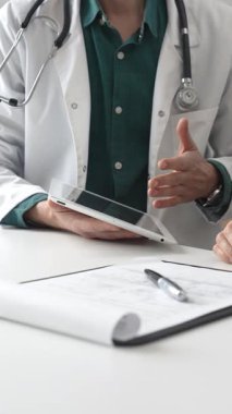 Yeşil gömlekli bir erkek doktor ve modern tıp ofisinde tablet kullanan hastaya teşhisi açıklarken steteskop hareketi yapıyor. İlaç ve sağlık hizmetleri kavramı.